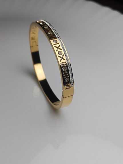 Gold Plated Roman Numeral Pave Crystal Bangle | Stainless Steel Hinged BraceletIntroduce a touch of sophisticated glamour to your collection with this stunning 24K Gold Plated Roman Numeral Bangle. This contemporary classic seamlessly blends luxurious design with everyday durability. The high-shine gold plating highlights the intricately engraved Roman numerals, which are beautifully accented by a continuous row of sparkling pave crystals along the edge, catching the light with every movement. Designed with a secure, hinged closure, this bracelet is easy to wear and stays comfortably in place. Crafted from premium stainless steel, it promises long-lasting shine and resistance to tarnish, making it the perfect statement piece for any occasion—from a casual brunch to a formal evening event. Key Features: Premium Material: Durable and tarnish-resistant stainless steel base. Luxe Finish: Rich, high-quality 24K gold plating for a lasting, brilliant shine. Elegant Design: Engraved with iconic Roman numerals for a sophisticated, classic look. Sparkling Accents: Adorned with a continuous line of brilliant pave crystals. Comfort & Fit: Features a secure hinged closure for easy on-and-off. Perfect Gift: An ideal gift for birthdays, anniversaries, or special occasions. Style: Stackable design—perfect for layering with other bracelets or wearing solo as a statement piece.